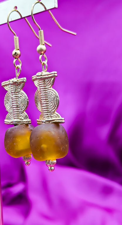Ces boucles d'oreilles réalisées  composées d'une perle translucide brunes ainsi qu'une perle doré ( poids Akan ) en laiton. 